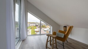 Habitación espaciosa con techos altos, ventanas amplias, suelo de madera, una única silla naranja y un taburete pequeño de madera.