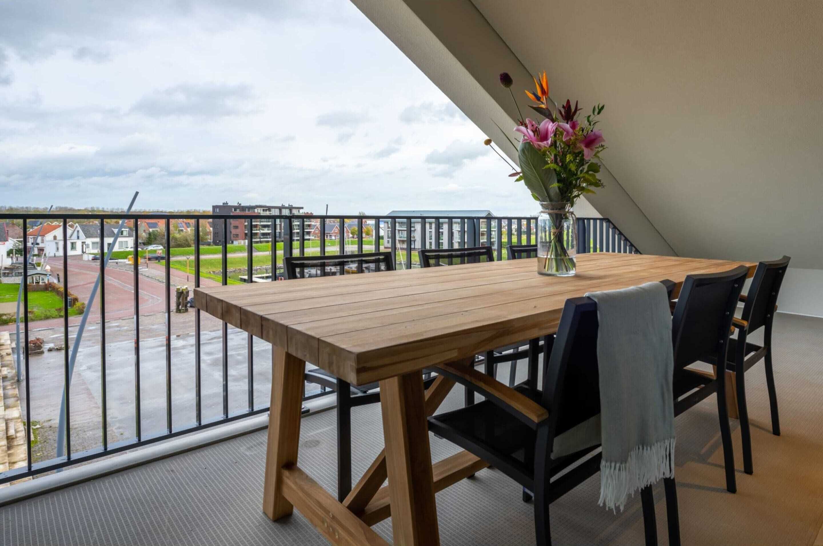 Ett träbord med stolar, en vas med blommor och en utsikt över husen genom en balkongräcke.