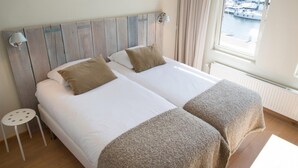 A double bed with two pillows, a small side table, and a view of a marina through a window. - Modern Apartment Overlooking the Harbor (The Hague)