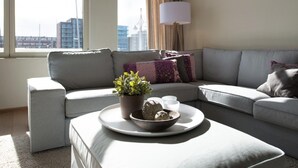 A modern living room with a grey sofa, a small side table with a plant and decorative items, and a view of the city through large windows. - Modern Apartment Overlooking the Harbor (The Hague)