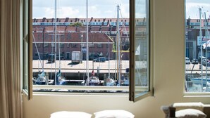 A room with a large window offering a view of a marina with boats and a building with red roofs. - Modern Apartment Overlooking the Harbor (The Hague)