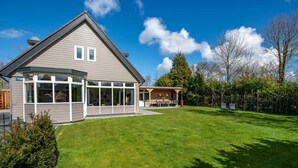 A modern house with a large lawn, a covered patio, and a separate building with a chimney. - Detached new 10-person Villa in Holiday Park (Kamperland)