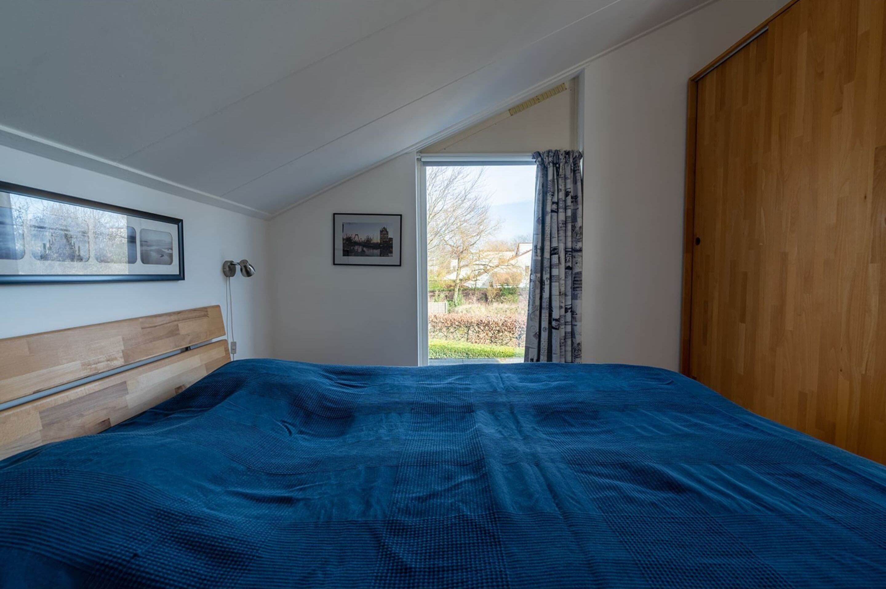 A bedroom with a wooden headboard, a large bed with a blue bedspread, a window with a view of greenery, and a wooden door.
