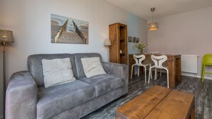 A modern living room with a grey sofa, wooden coffee table, and a dining area with white chairs and a wooden table. - Holiday Home Near the Beach Zoutelande (Zoutelande)