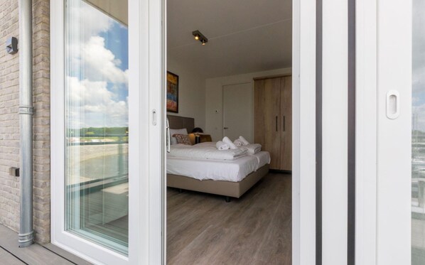 A bedroom with a bed, a nightstand, a lamp, and a view of the outdoors through a glass door. - Apartment in Sint-annaland by Marina (Sint-Annaland)