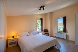A bedroom with a bed, nightstand, chair, and two windows with curtains. - Bungalow Near Oosterschelde Beach (Kamperland)