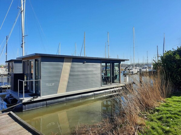 Un barco moderno de un solo nivel con fachada de vidrio, llamado 'TULIP', atracado en un puerto deportivo con varios veleros al fondo.