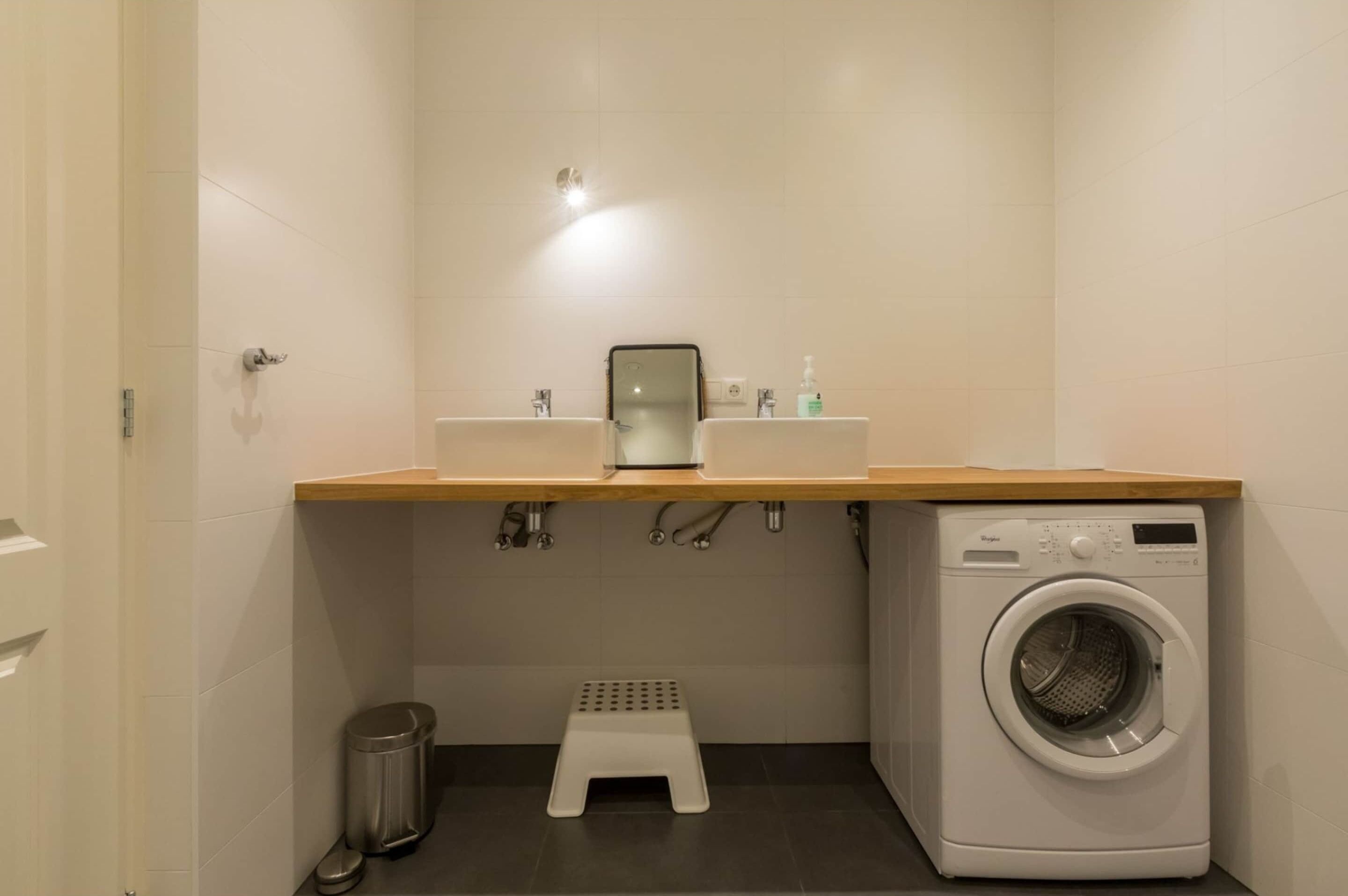 A compact bathroom with a washing machine, a small step stool, and a trash can.