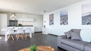 Una sala de estar moderna con un sofá gris, una mesa de centro de madera y un comedor con sillas blancas, con una cocina al fondo.