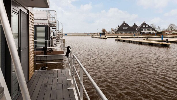 A boat deck with a 'SLOW MOTION' sign, wooden flooring, metal railings, and a view of houses across the water. - Houseboat With Stunning Views (Offingawier)