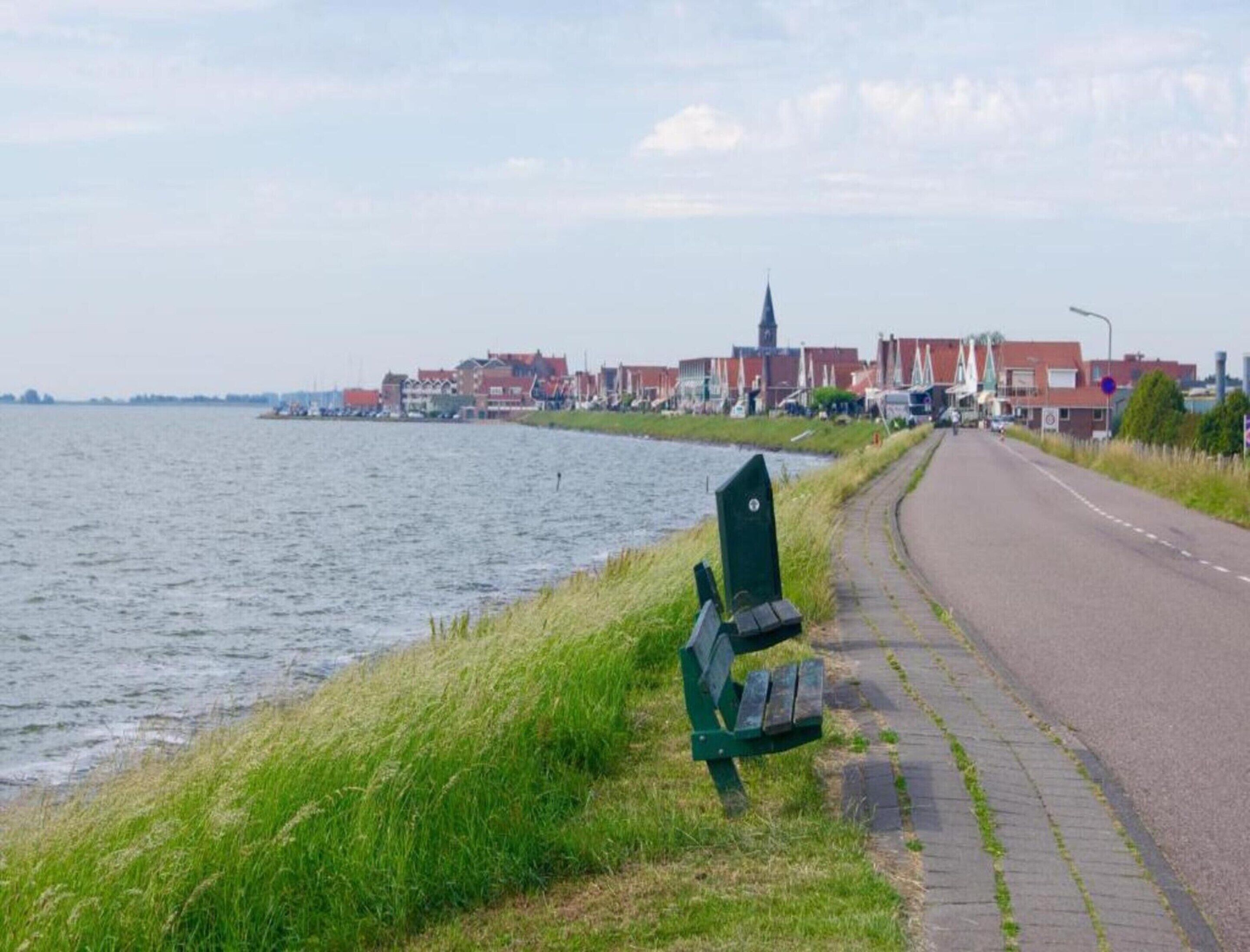 A riverside walkway with benches, a road, and buildings in the background.