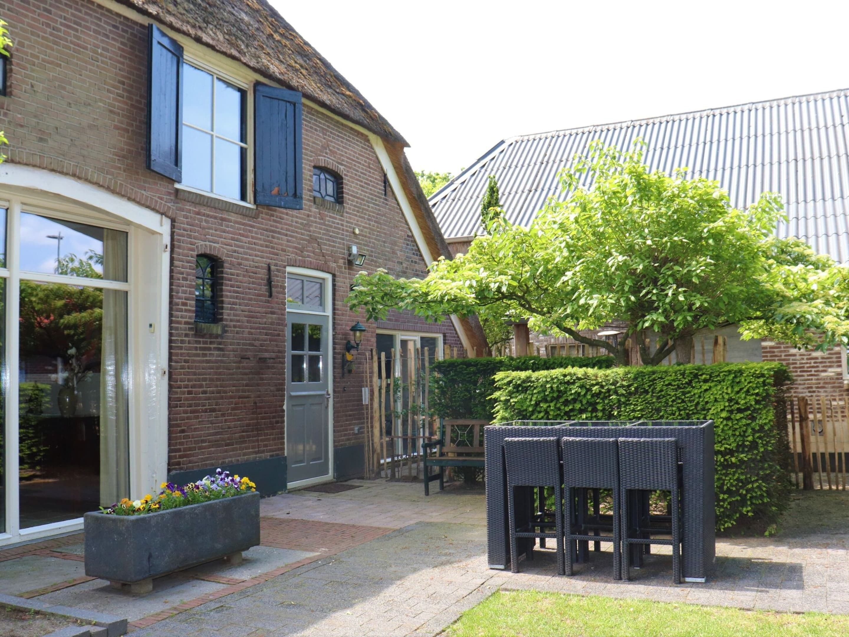 Et murhus med stråtækt tag, en terrasse med fletningsmøbler og en stor plantekasse med blomster.