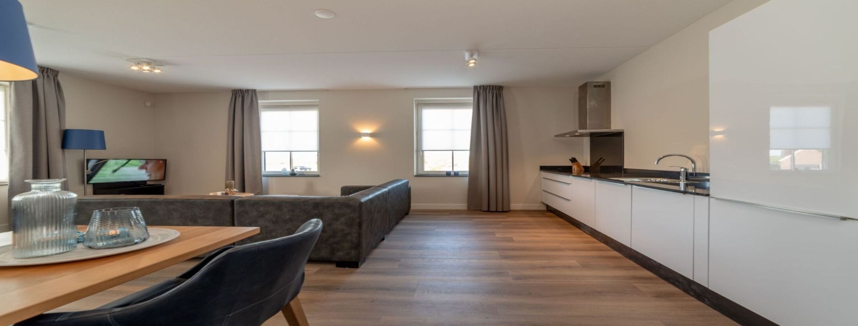 A modern apartment with a dining area, a living room with a sofa, a kitchen with white cabinets, and a large window providing natural light.