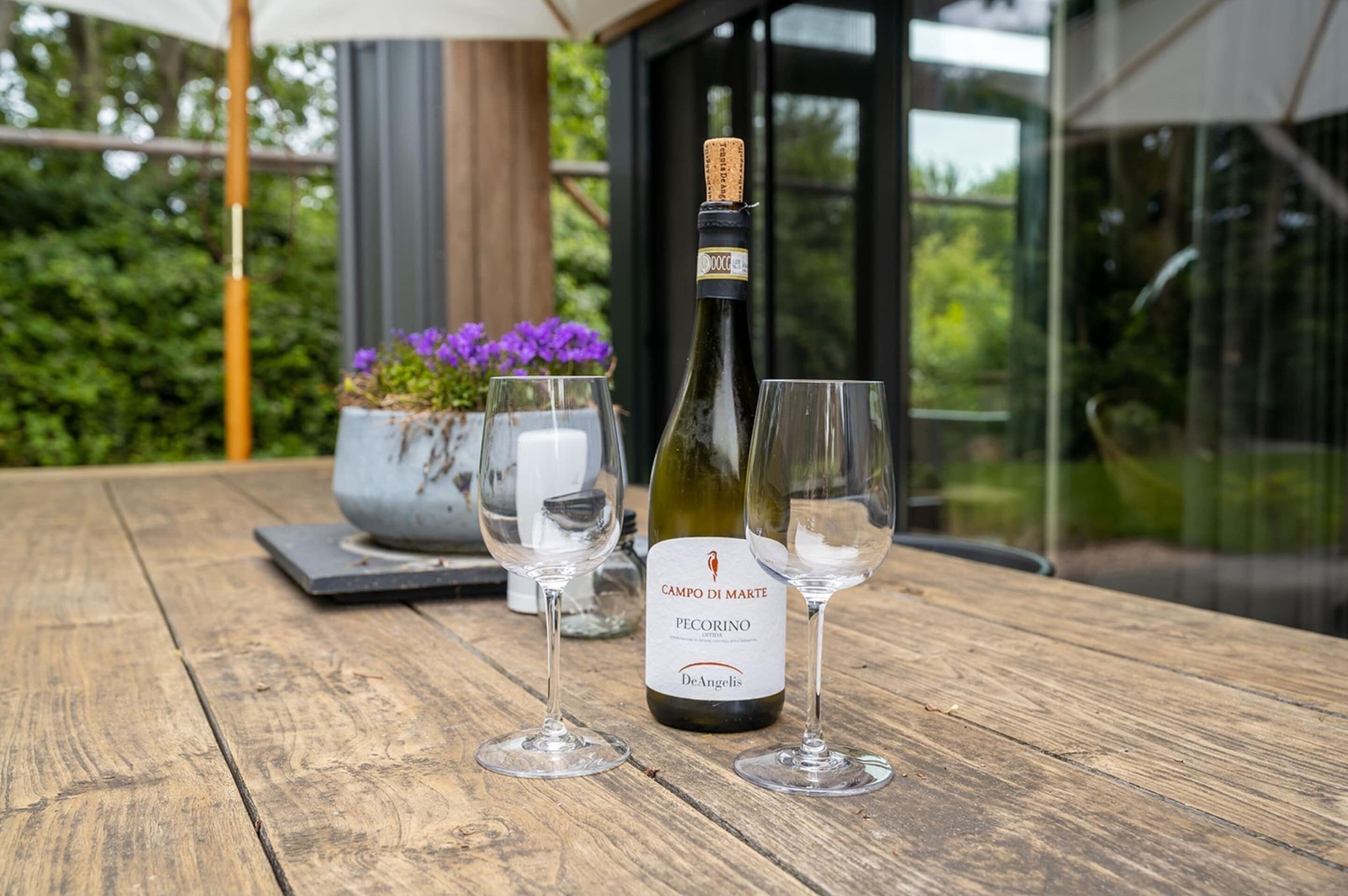 A bottle of wine and two glasses on a wooden table with a potted plant and a white umbrella in the background.