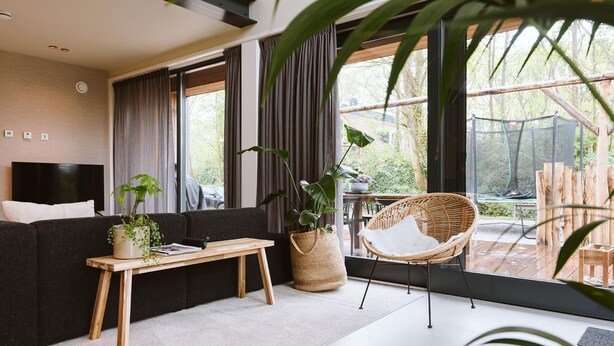 Una sala de estar moderna con un sofá, una mesa de centro de madera, una silla de mimbre y ventanas amplias con vistas al exterior.