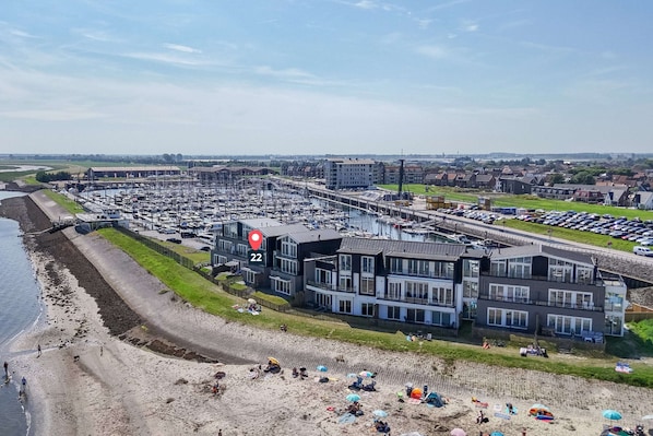 Een jachthaven met boten, een strand en een modern gebouw met meerdere verdiepingen en grote ramen.