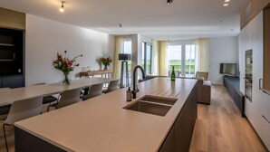 A modern kitchen with a central island, a dining area with chairs, a TV, and a balcony with a view.