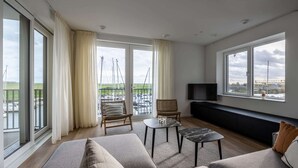 A spacious living room with a large window, a sofa, a small table, and a TV mounted on the wall.