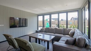 A spacious living room with a large sectional sofa, a flat-screen TV mounted on the wall, and a dining table with chairs. - Attractive Holiday Home in Callantsoog With Fenced Garden (Callantsoog)