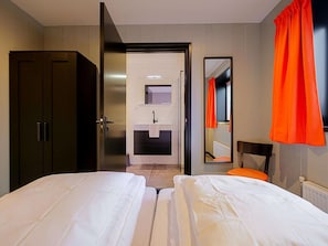 A hotel room with two beds, a dark wardrobe, a bathroom with a sink and mirror, and orange curtains.