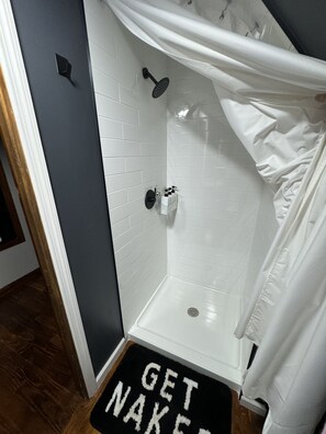 A bathroom with white tiled walls, a shower with a black handheld showerhead, and a white shower curtain. There is a black mat with white text at the entrance.