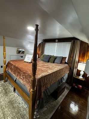 A bedroom with a four-poster bed, a bedside table with a lamp, a rug, and a window with curtains.