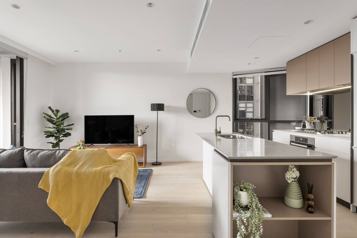A modern living room with a grey sofa, a yellow throw blanket, and a TV. The kitchen has a large island with a sink and a stove.