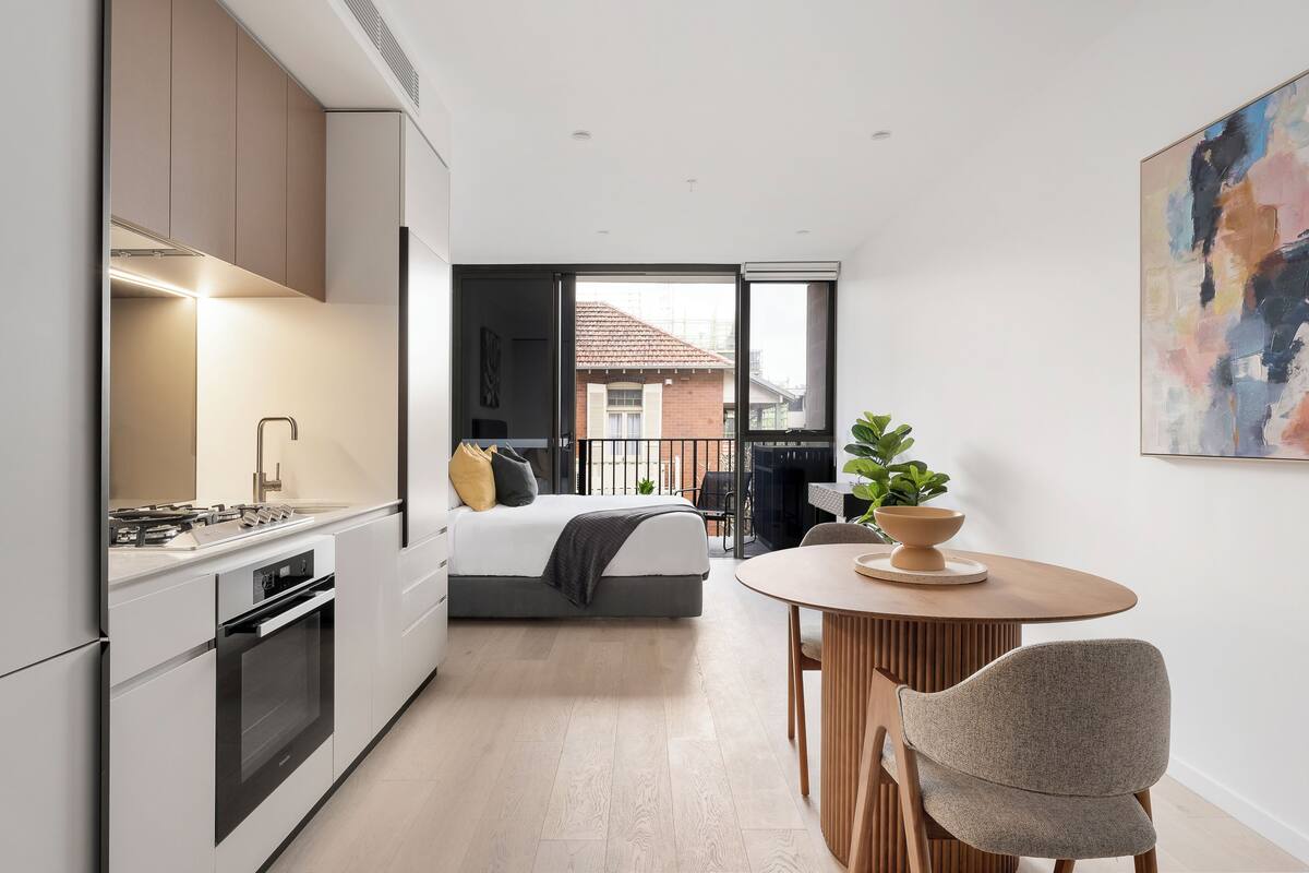 A modern kitchen with integrated appliances, a dining area with a round table and chairs, a bed with a view to the outside, and a large abstract painting on the wall.