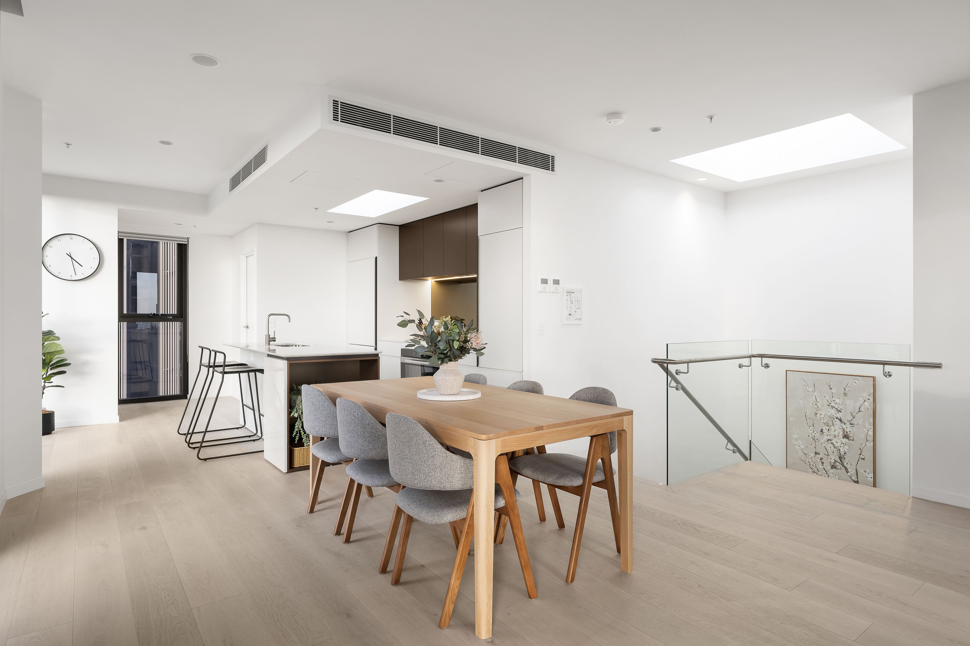 A modern kitchen with a wooden dining table, grey upholstered chairs, and a glass railing.