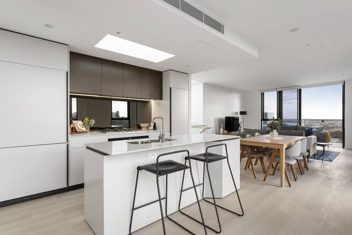 A modern kitchen with a central island, bar stools, a dining area with a wooden table and chairs, and large windows offering a city view.