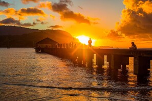 Une jetée s’avançant dans des eaux calmes au coucher du soleil, avec la silhouette de personnes et une structure sur la gauche.