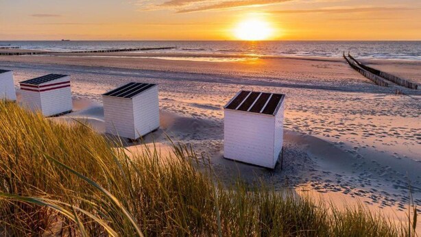 Eine Strandansicht mit Sonnenuntergang, mit drei weißen Gebäuden mit Solarpanels, einem Sandstrand und einem hölzernen Steg im Hintergrund.