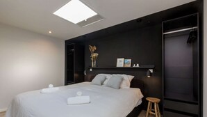 Ein modernes Schlafzimmer mit einem großen Bett, einem kleinen Hocker, einem dunklen Kopfteil und einem Regal mit Gegenständen.