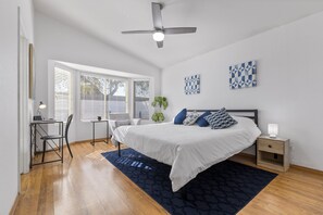 A bedroom with a bed, bedside table, lamp, chair, desk, and window with blinds.