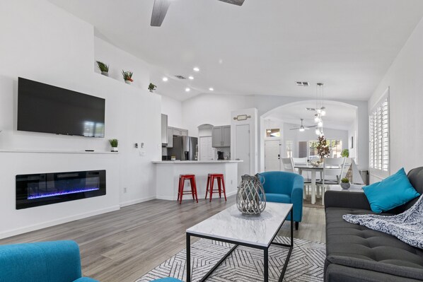 A modern living room with a flat-screen TV, a fireplace, a dining area with a table and chairs, and a kitchen in the background.