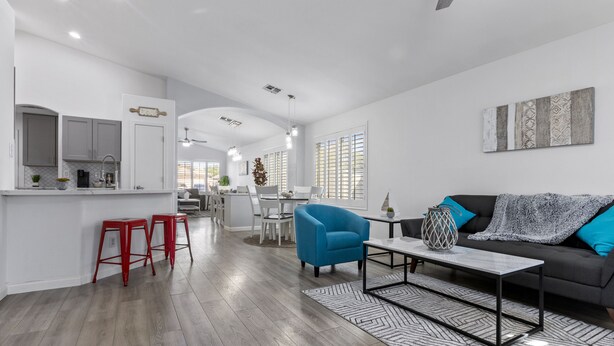 Una sala moderna con un sofá gris, un sillón azul y una mesa de centro. Cuenta con una cocina con armarios blancos y un comedor con una mesa y sillas blancas.