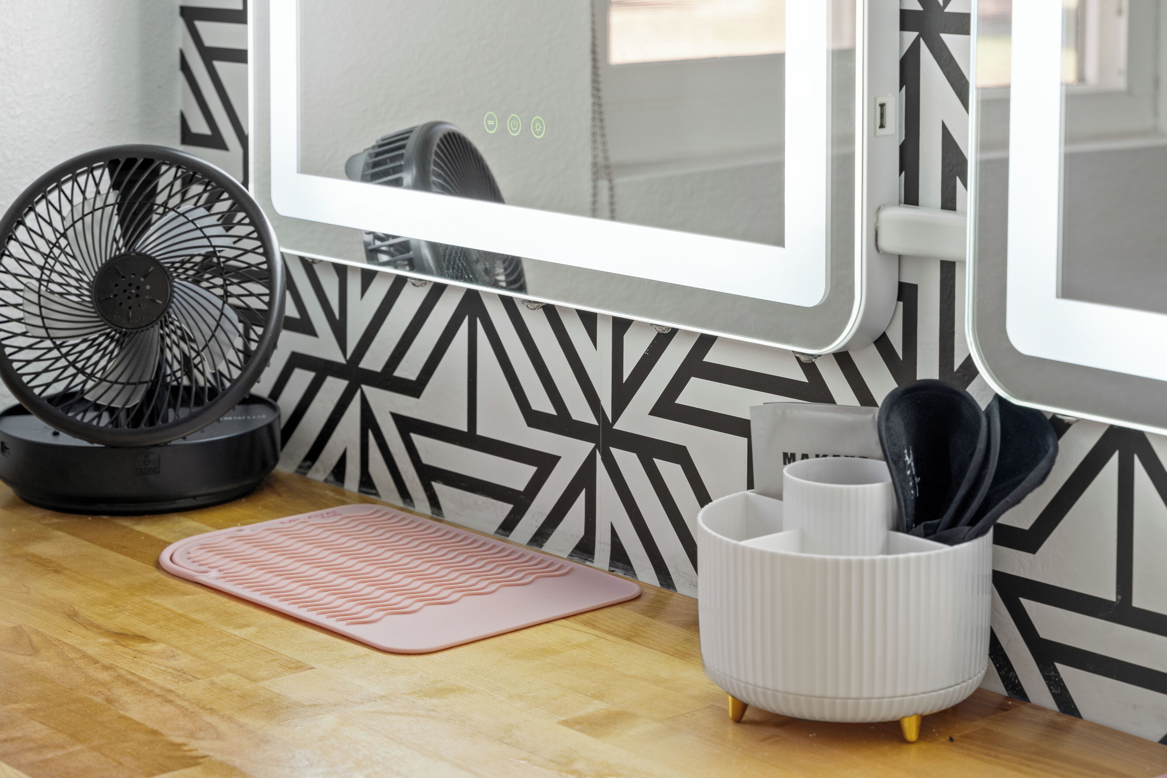 Una habitación con una mesa de madera, un ventilador, un portalvaques blanco y un tapete rosa.