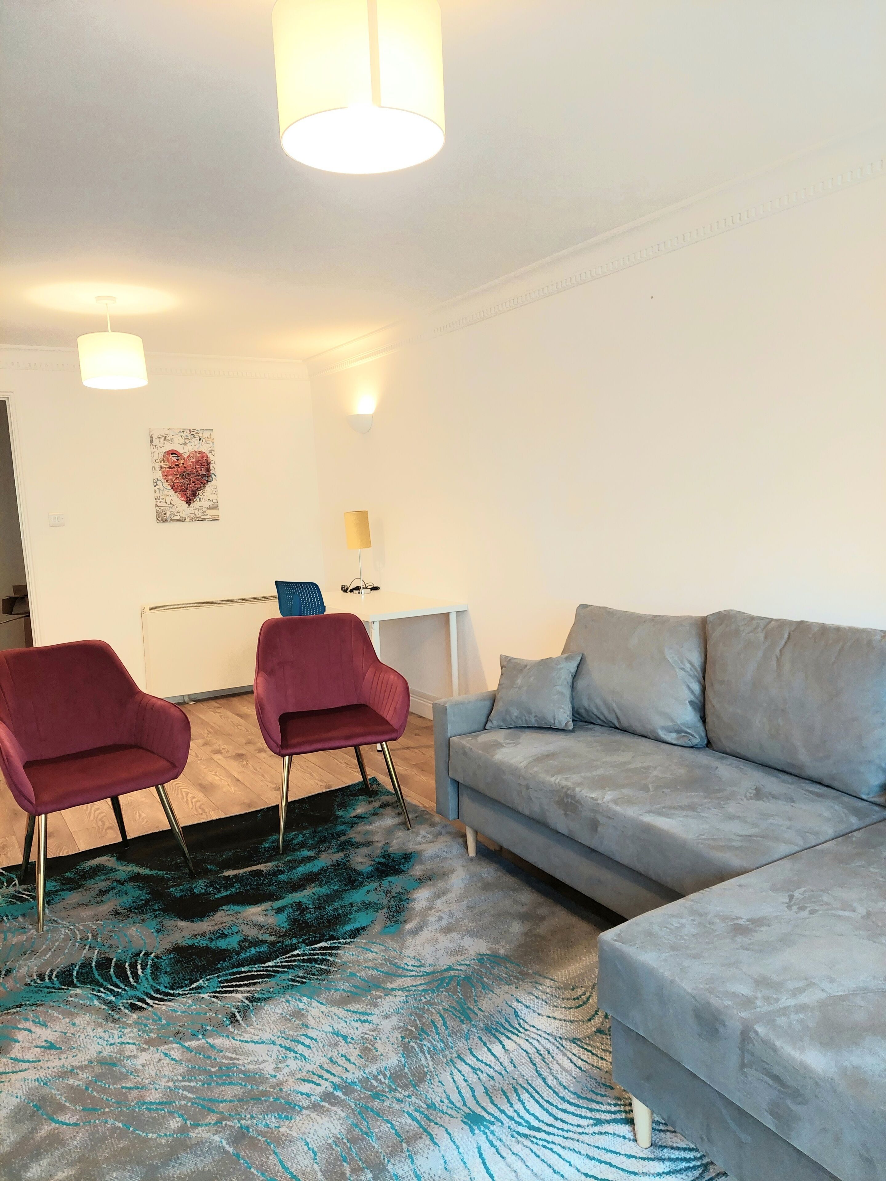 Een woonkamer met een grijze bank, twee rode stoelen, een kamerkleed met patroon en een muur met een hartvormig kunstwerk.