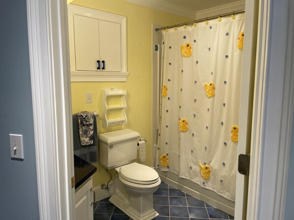 A bathroom with a toilet, a vanity with a patterned towel, a white cabinet above the toilet, and a shower curtain with duck designs. - Encounters at Lake Gaston - New (Bracey)