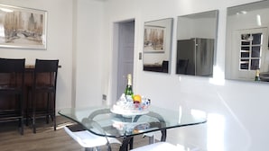 A dining area with a glass table, two chairs, and a bottle of champagne. There are framed paintings on the walls and a refrigerator in the background.