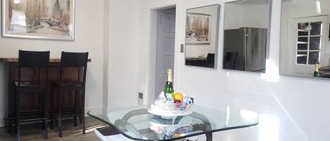 Un comedor con una mesa de vidrio, dos sillas y una botella de champán. Se observan cuadros enmarcados en las paredes y un refrigerador al fondo.