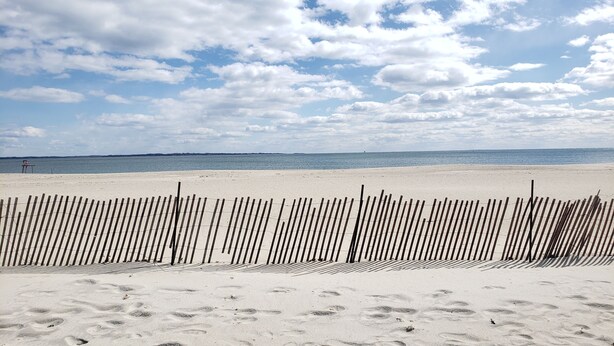 A sandy beach with a wooden fence in the foreground and a clear sky with clouds in the background.