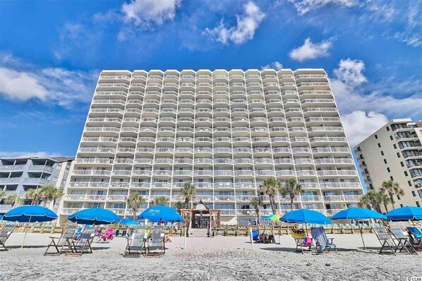 A large hotel with multiple balconies, a beach area with umbrellas, and palm trees. - Breathtaking Views Oceanfront 2 Bed 2 Bath Condo Water's Edge Resort Garden City (Murrells Inlet)