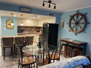 A compact kitchen with a dining area, featuring a round glass table, chairs, a black refrigerator, and a wooden ship's wheel decoration. - Breathtaking Views Oceanfront 2 Bed 2 Bath Condo Water's Edge Resort Garden City (Murrells Inlet)