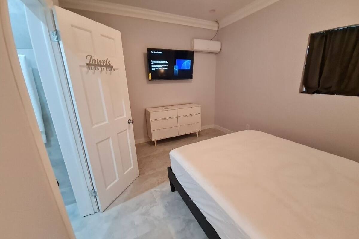 A bedroom with a bed, a TV mounted on the wall, a dresser, and a door with a towel rack.