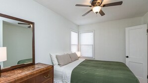 Une chambre à coucher comprenant un lit, une commode, un miroir, un ventilateur de plafond et deux fenêtres.
