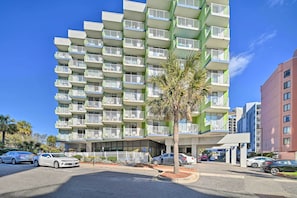 A multi-story hotel with balconies, a palm tree, and cars parked in front.