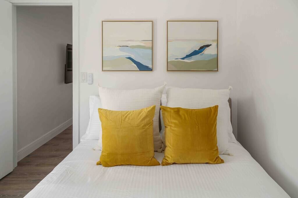 Habitación de hotel con cama, ropa de cama blanca, dos almohadas amarillas y dos cuadros abstractos en la pared.