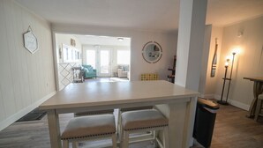 Un área de comedor con una mesa de color claro y cuatro taburetes. Hay un espejo en la pared y una lámpara de pie a la derecha.