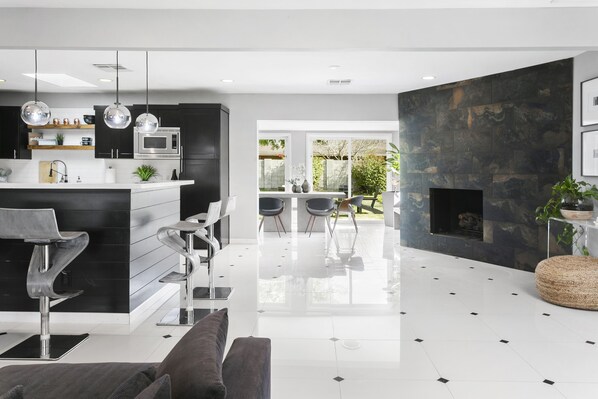 A modern kitchen with black cabinets, stainless steel appliances, and a central island. There is a dining area with a table and chairs, and a fireplace with a stone wall. - POOL▪️FIREPITS▪️CORNHOLE▪️1/3 ACRE LOT (Phoenix)